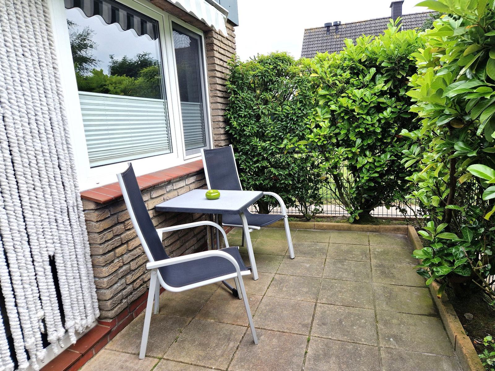Terrasse mit Tisch und zwei Stühlen, umgeben von grünen Sträuchern und einem Haus.