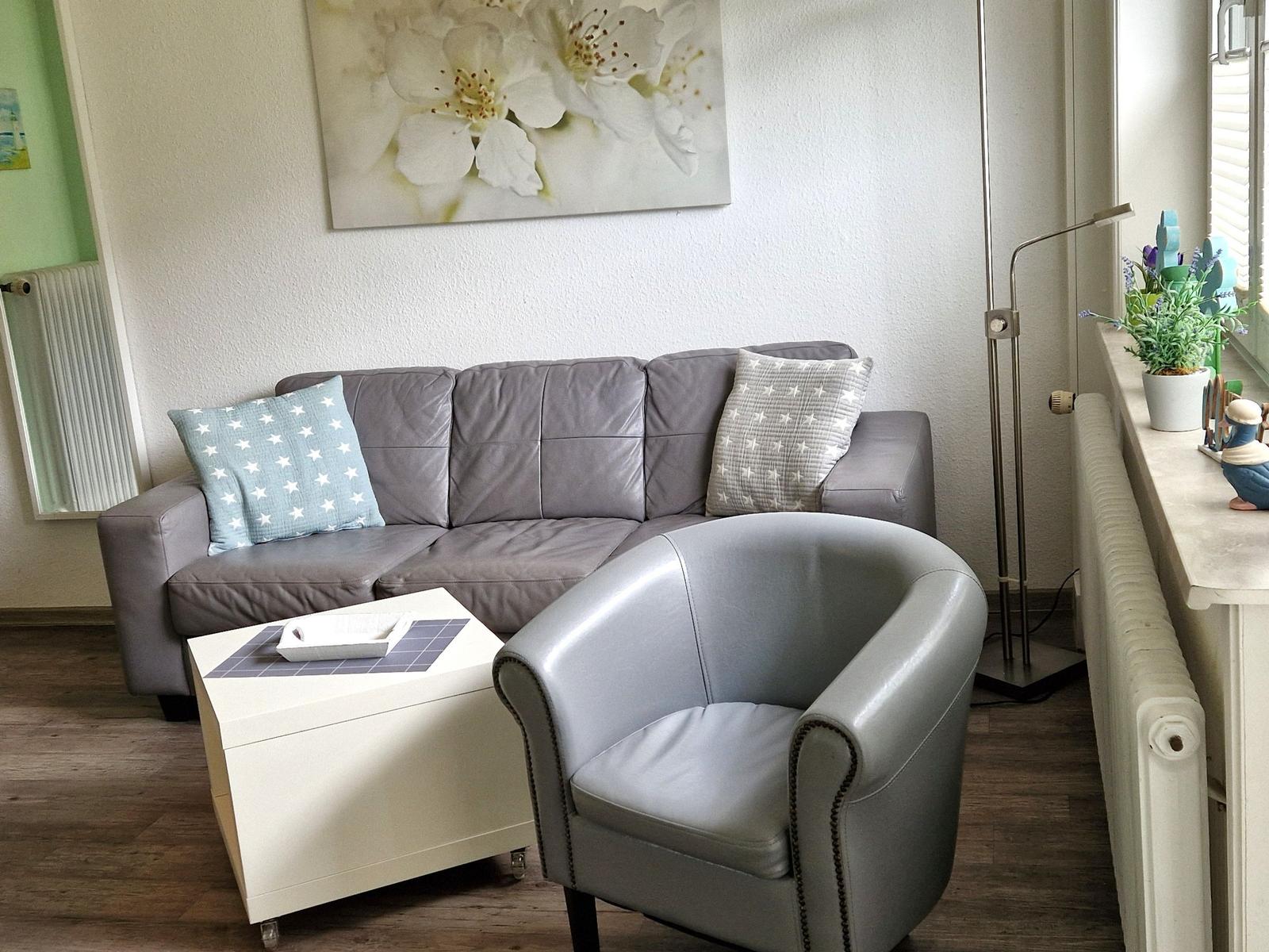 Ein Wohnzimmer mit grauem Sofa, Stuhl und Couchtisch. An der Wand hängt ein Bild mit Blumen.