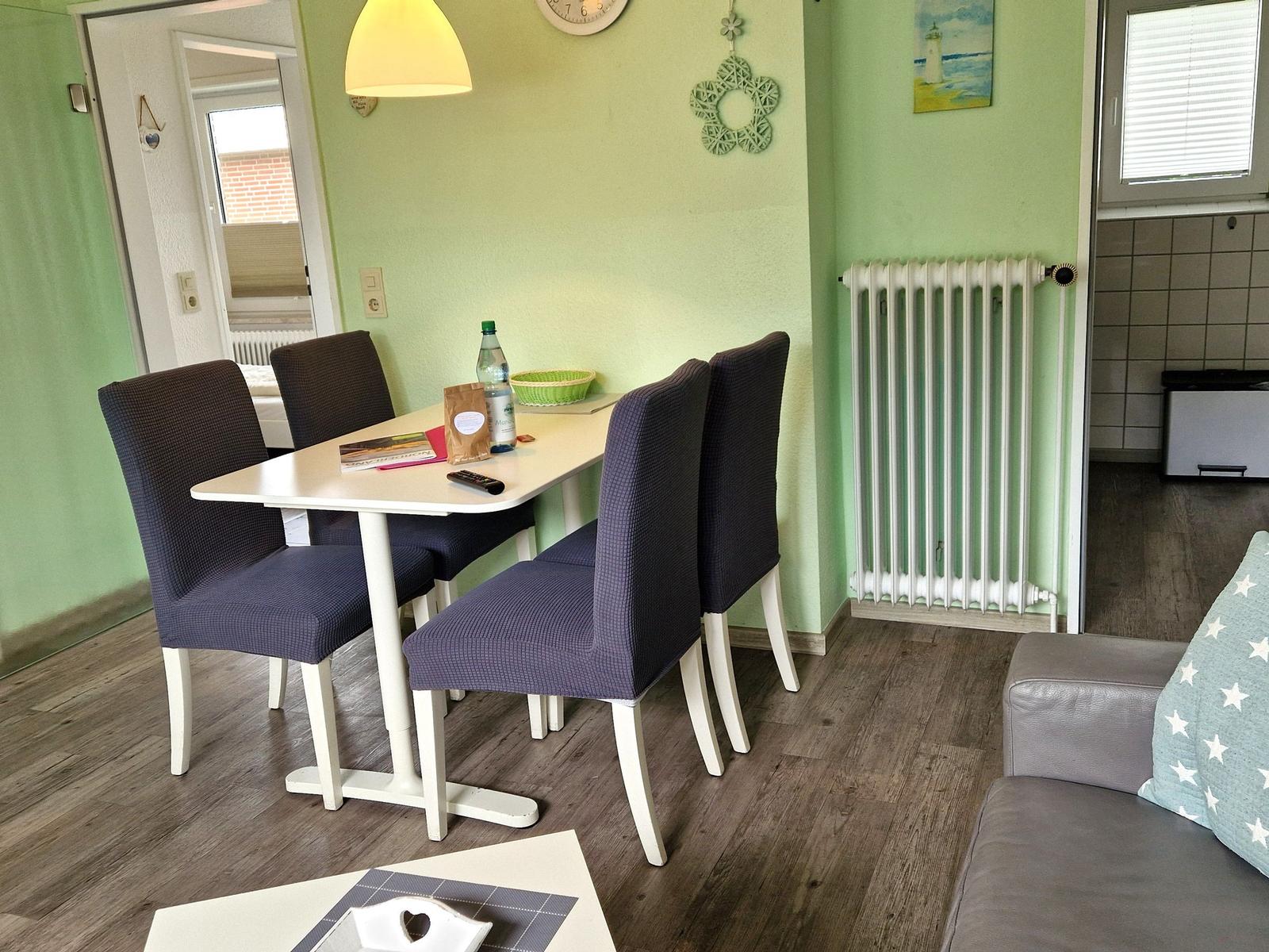 Esszimmer mit Tisch, vier Stühlen und grünen Wänden. Sofa und Heizung sind sichtbar.