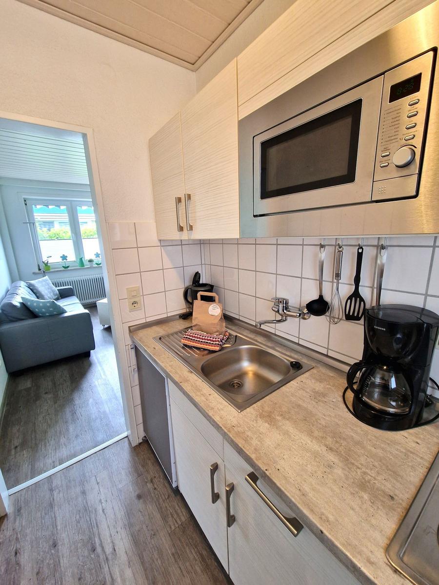 Küche mit Spüle, Mikrowelle und Kaffeemaschine. Wohnzimmer mit Sofa und Fenster sichtbar.