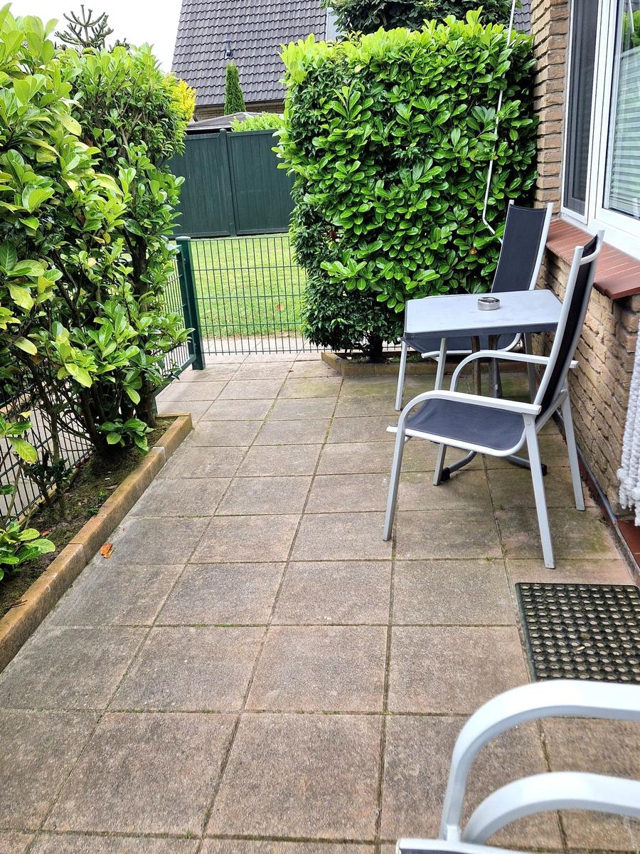 Terrasse mit Tisch, zwei Stühlen und grünen Hecken. Hinterhof mit Zaun und Rasen.