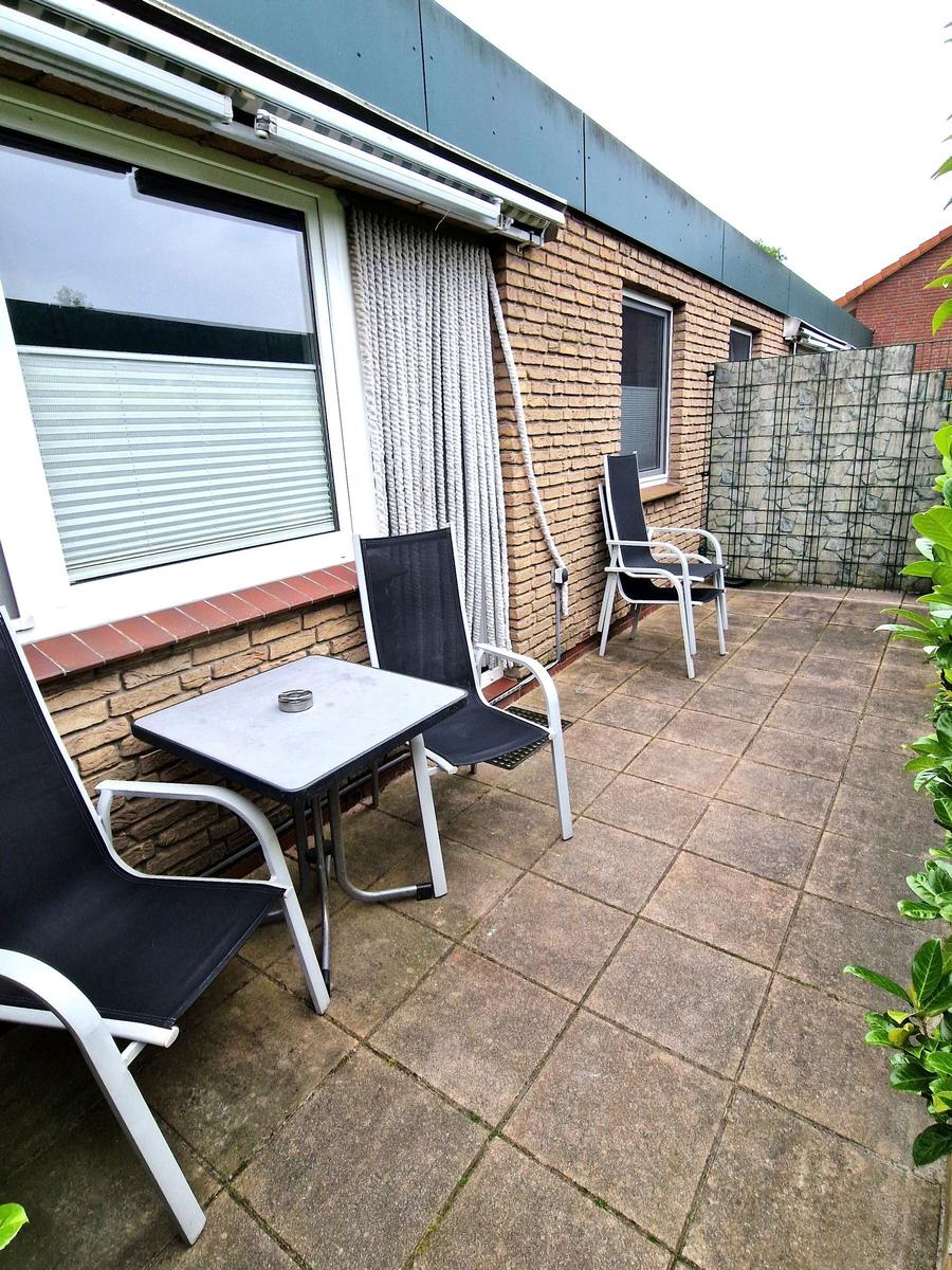 Terrasse mit Tisch und drei Stühlen, Wand mit Jalousie und Steinmauer im Hintergrund.