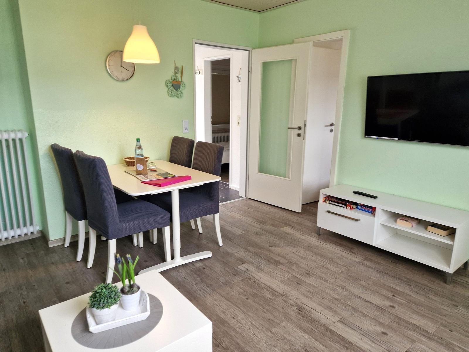 Wohnzimmer mit Esstisch, Fernseher und Tür zum Schlafzimmer.