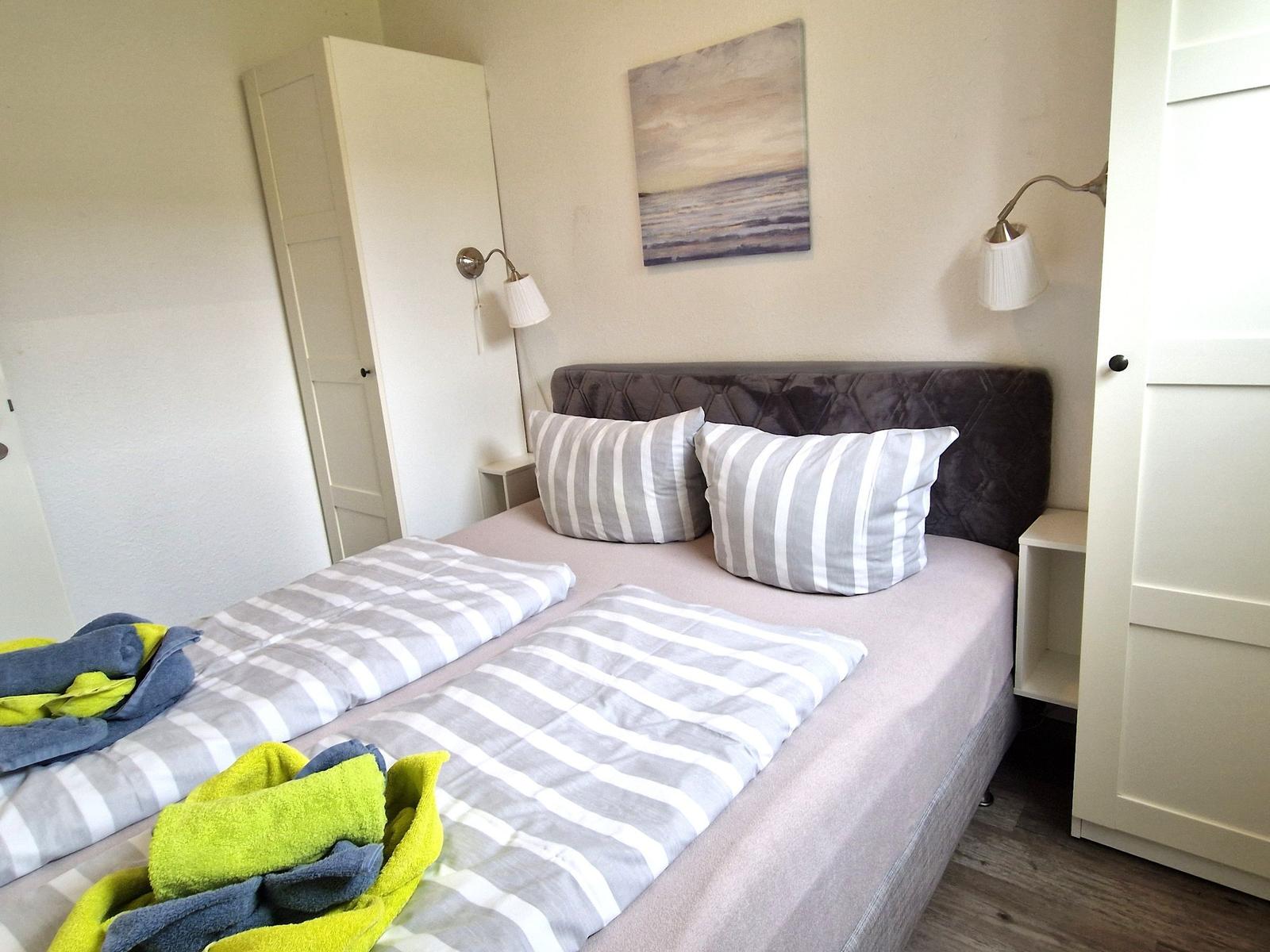 Schlafzimmer mit Bett, Kissen und Wandbild. Zwei Stehlampen und Schrank sind sichtbar.