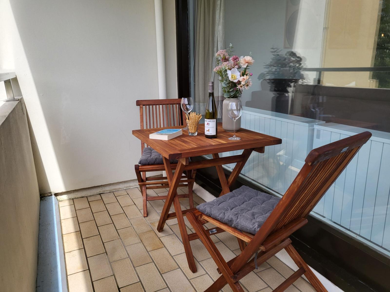 Terrasse mit Holztisch, zwei Stühlen, Blumen und Weinflasche.