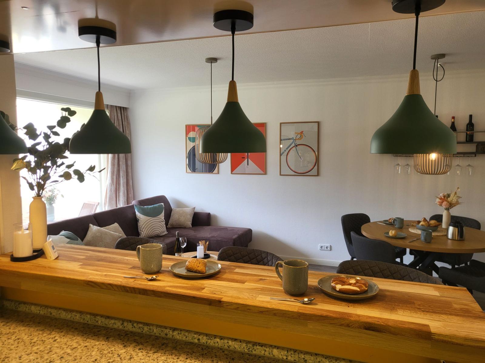 Essbereich mit Holztisch, Sitzgelegenheiten und Hängelampen. Wandbilder und Pflanzen sichtbar.