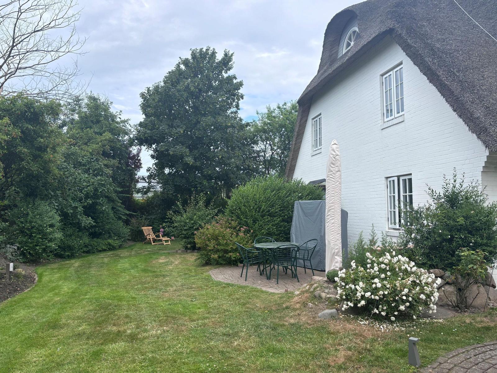 Haus mit strohgedecktem Dach und Garten mit Tisch, Stühlen und Blumen.