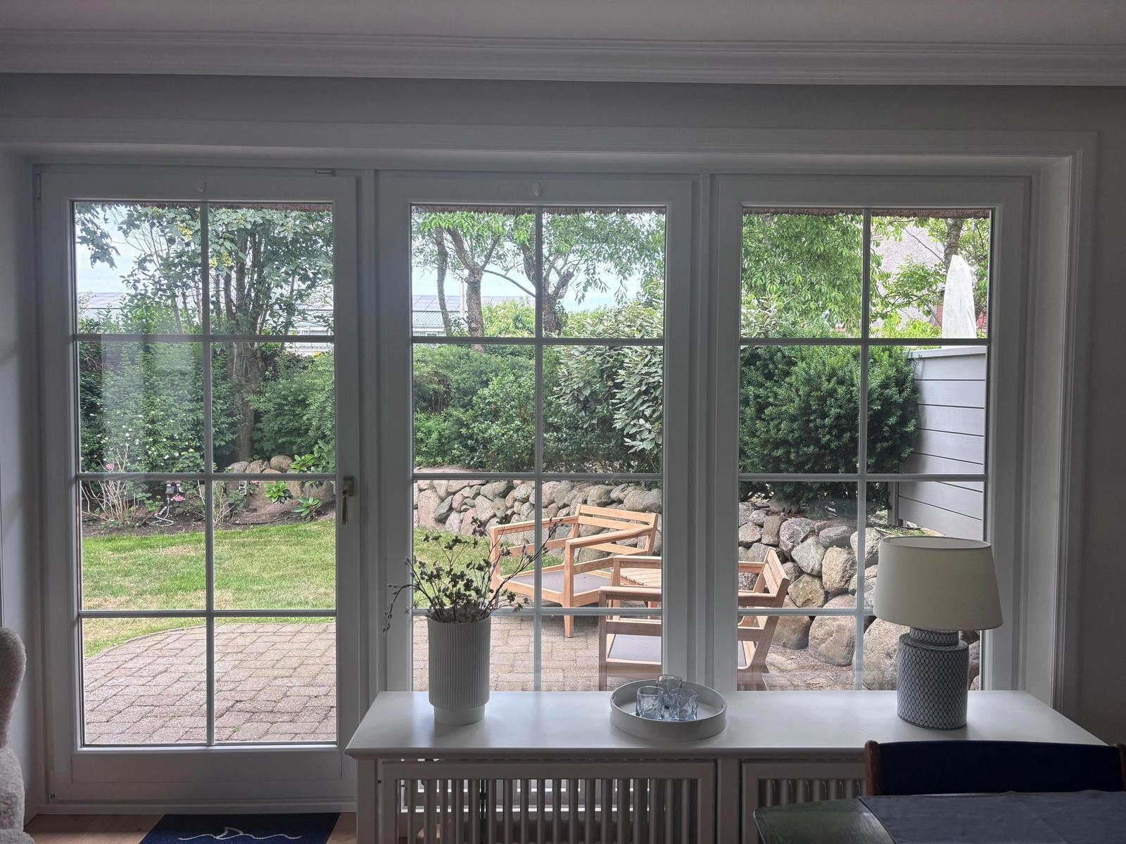 Durch große Fenster blickt man auf einen gepflegten Garten mit Holzstühlen und Steinmauer.