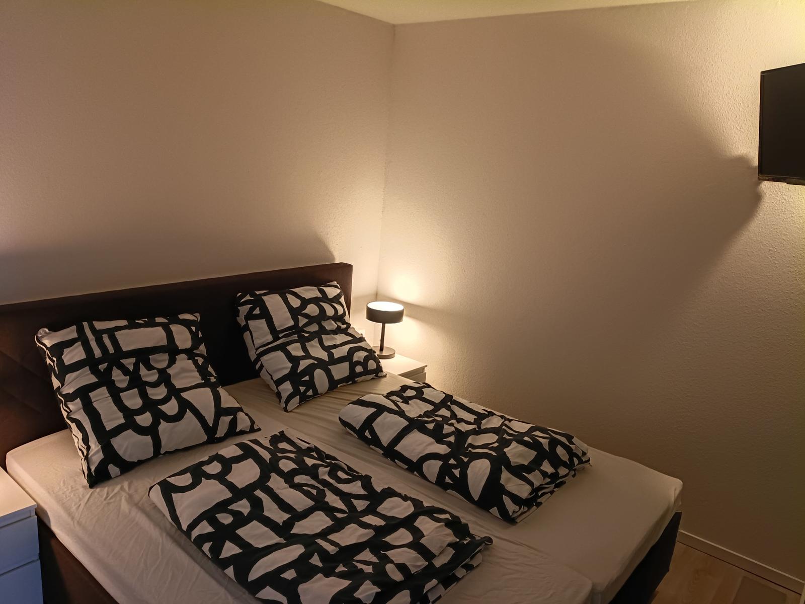 Schlafzimmer mit Bett, Kissen und Decke mit schwarz-weißem Muster, Nachttisch und Wand-TV.