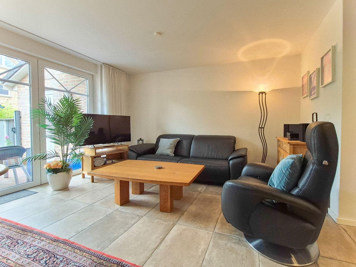 Wohnzimmer mit Sofa, Sessel, Tisch und Fernseher. Durchgang zur Terrasse.