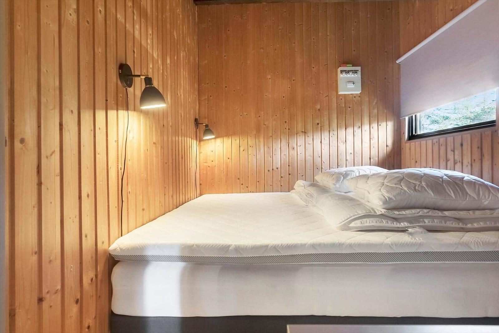 Schlafzimmer mit Bett, Holzwand und Wandlampen. Fenster mit Rollladen.