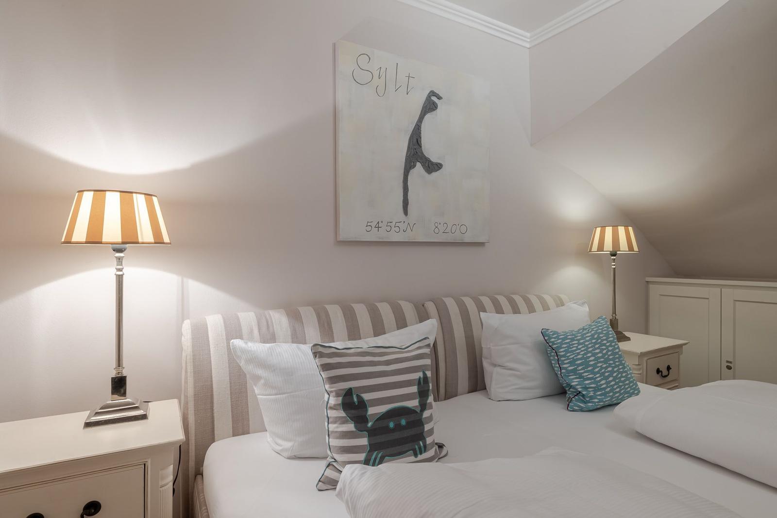 Schlafzimmer mit Bett, zwei Nachttischen und Lampen. Wandbild zeigt Sylt mit Koordinaten.