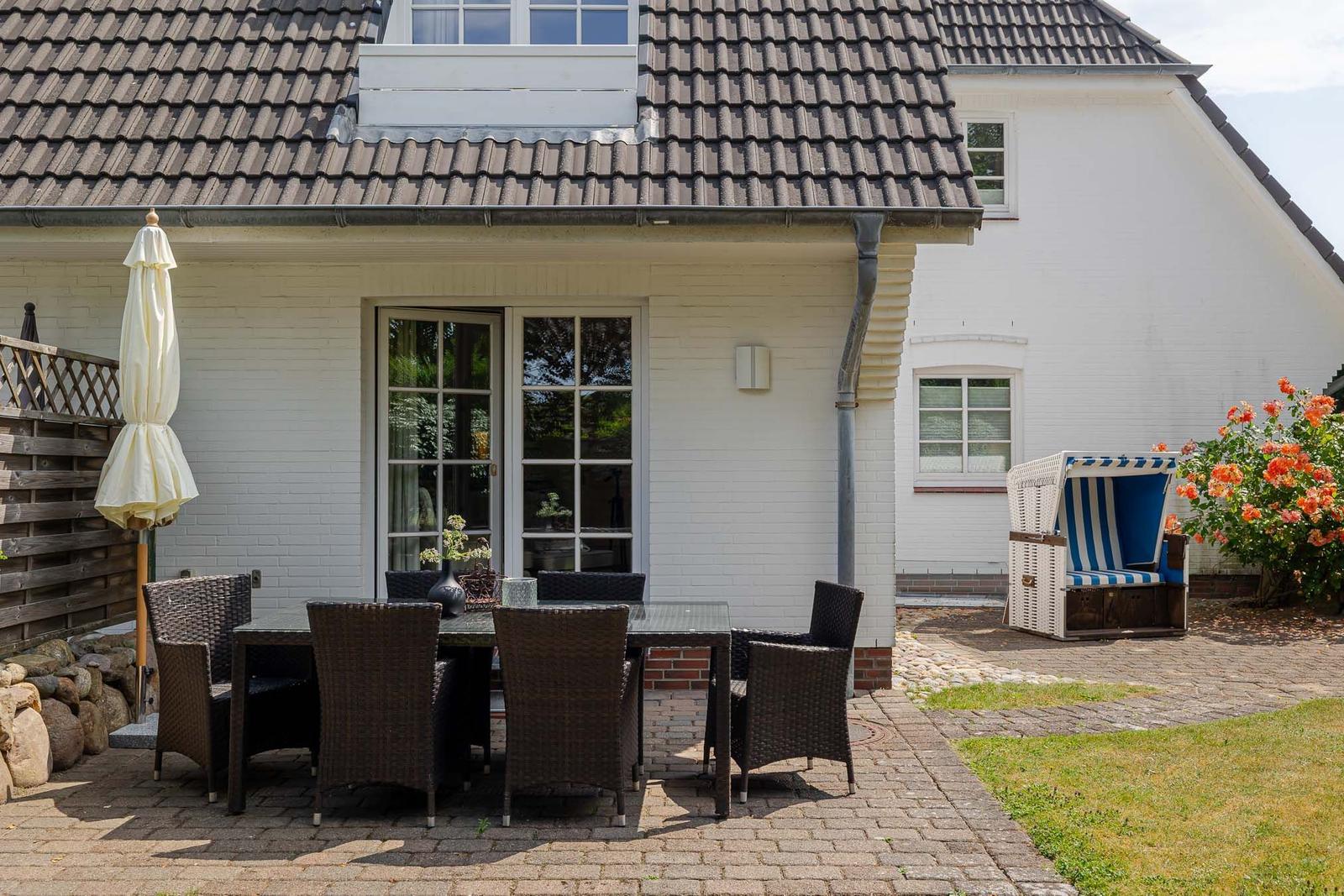 Terrasse mit Tisch, Stühlen und Liegestuhl. Hintergrund: weißes Haus mit Dach.