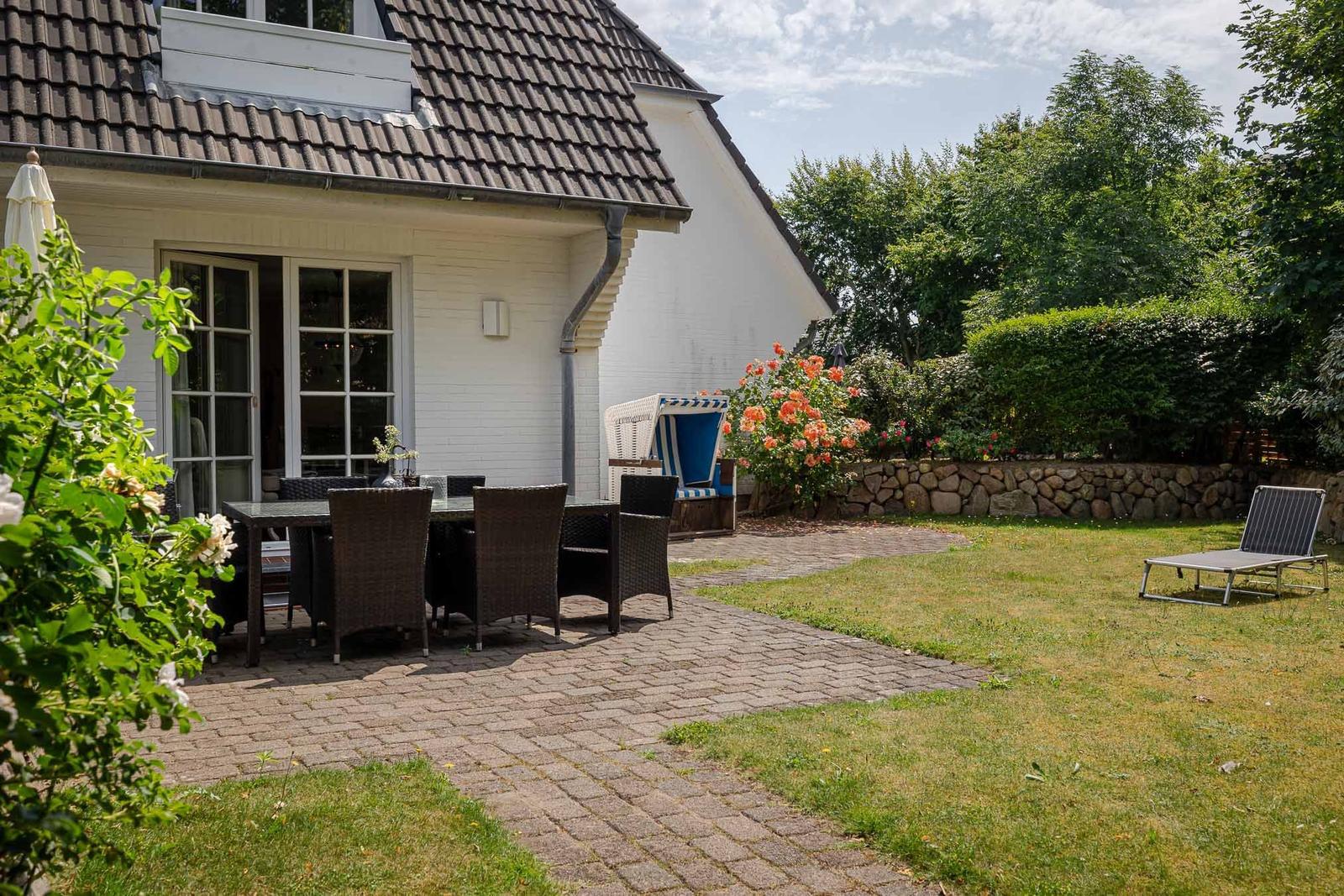 Haus mit Terrasse, Gartentisch, Liegestuhl und Blumenbeet.