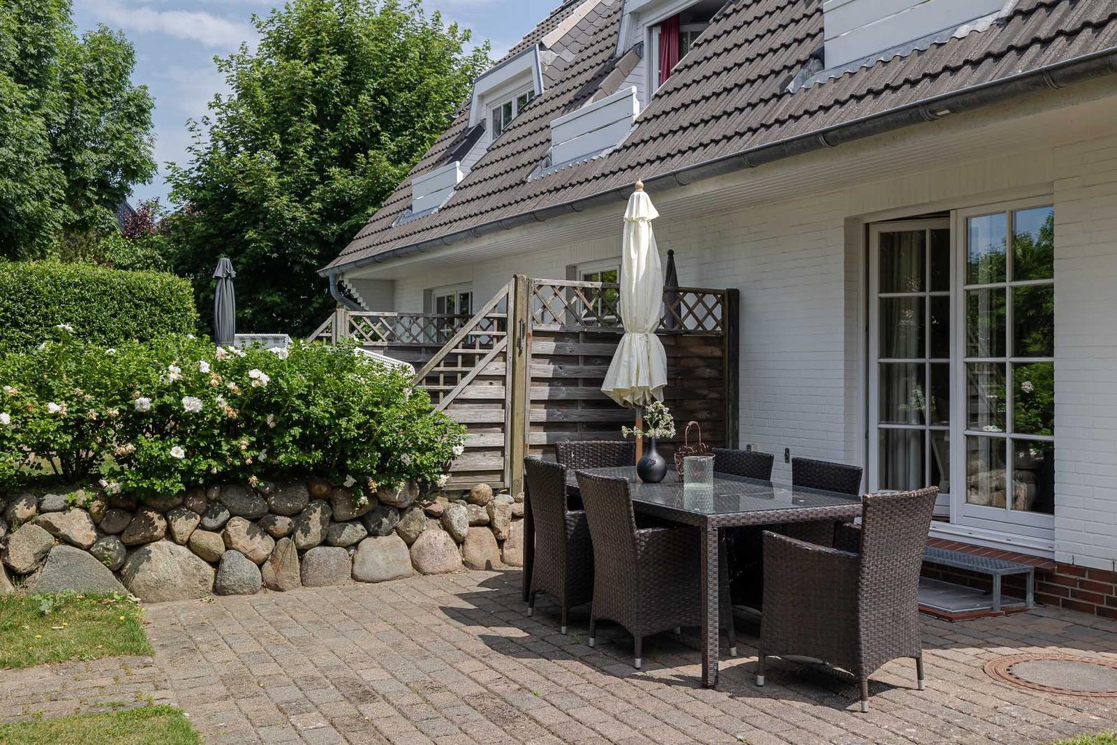 Terrasse mit Tisch, Stühlen und Sonnenschirm neben weißem Haus mit Steinmauer und Blumenbeet.