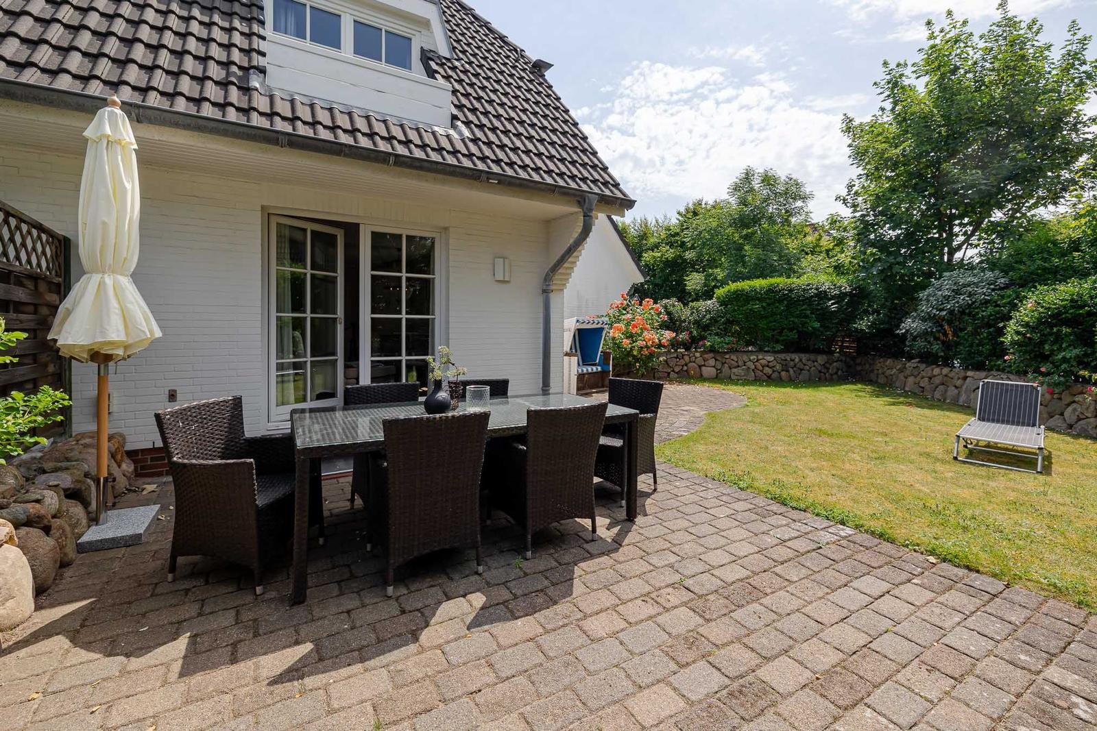 Hinterhof mit Terrasse, Tisch, Stühlen und Sonnenschirm. Grasfläche und Bäume im Hintergrund.