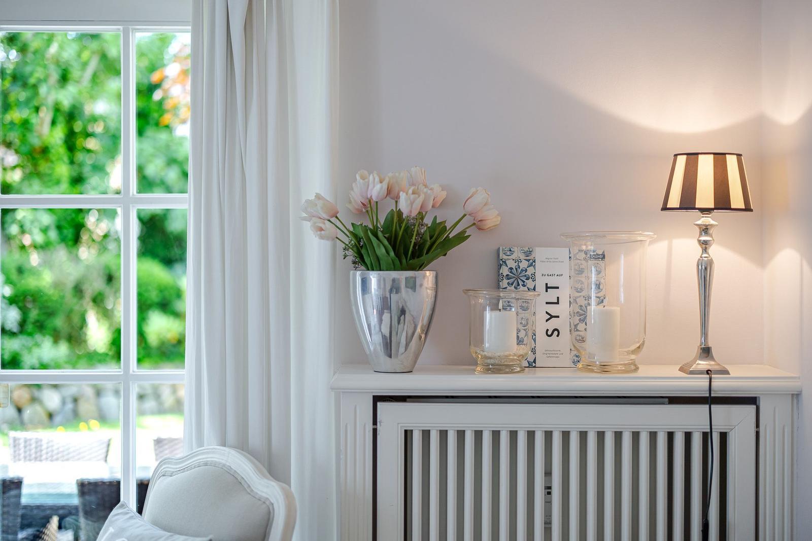 Ein Fenster mit Blick auf einen Garten. Auf einem Schrank stehen Blumen, Kerzen und eine Lampe.