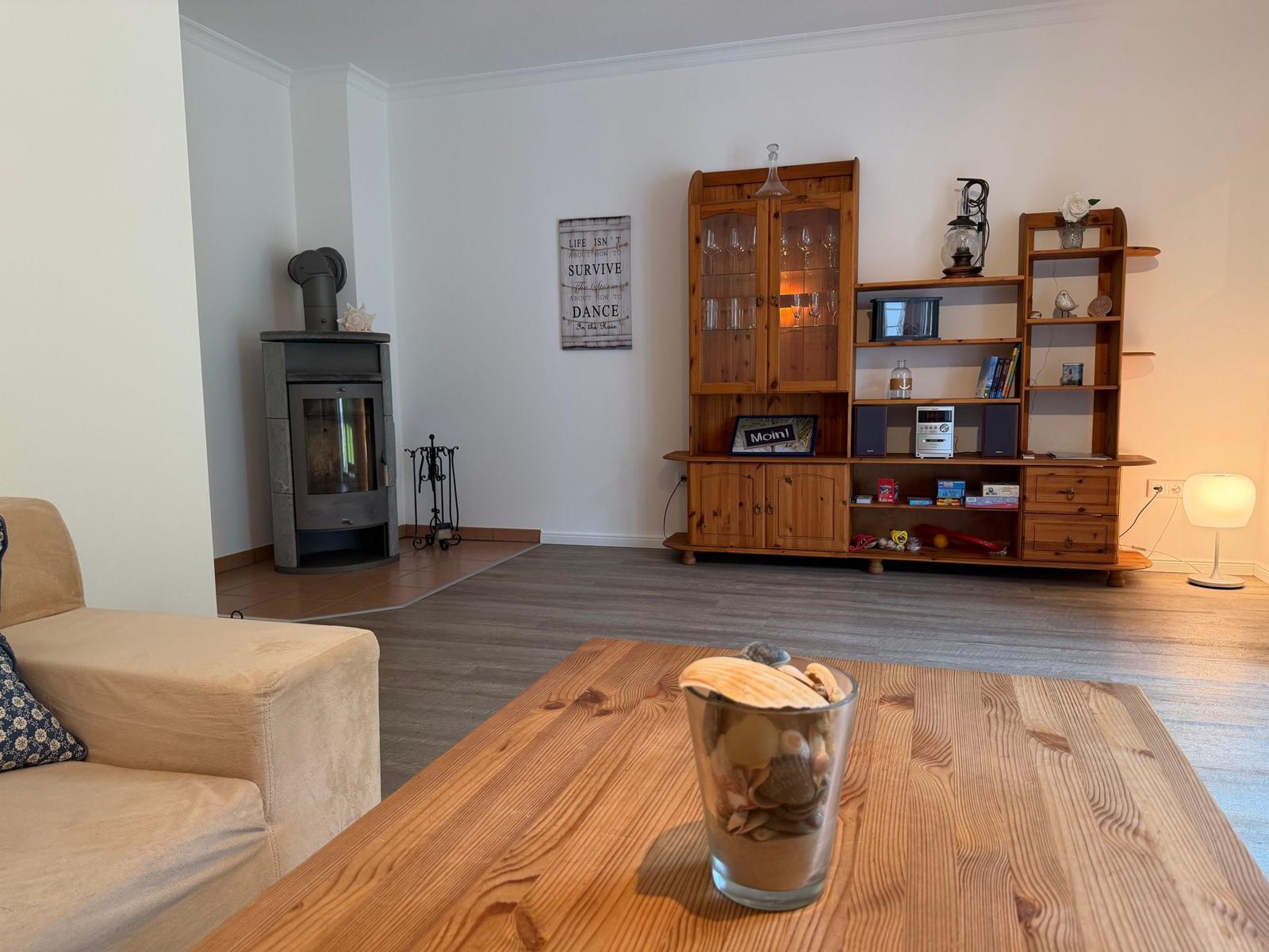 Wohnzimmer mit Holzregal, Kaminofen und Holztisch. Auf dem Tisch steht ein Glas mit Muscheln.