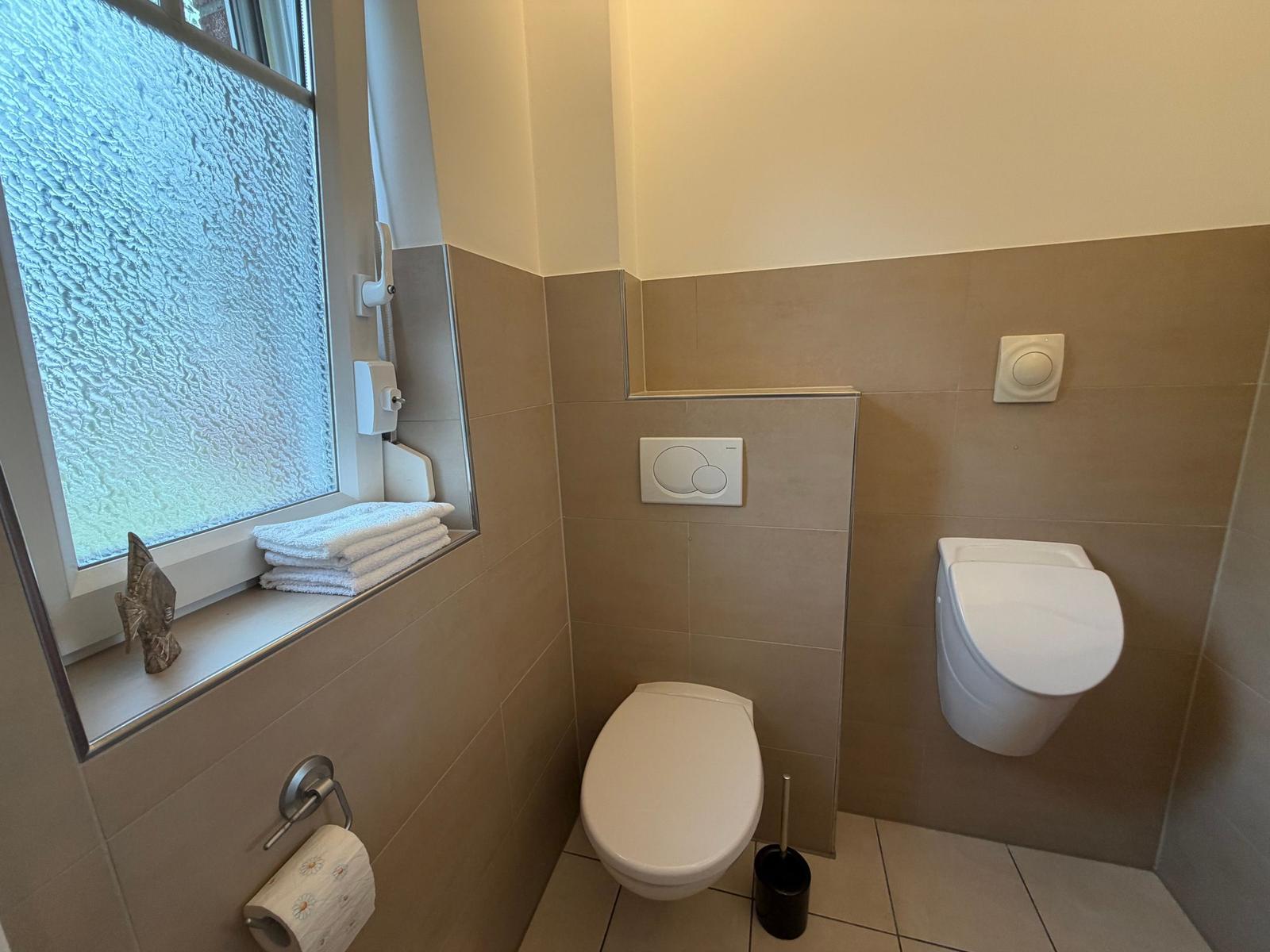 Badezimmer mit Toilette, Wandspüler und Fenster mit Handtüchern.