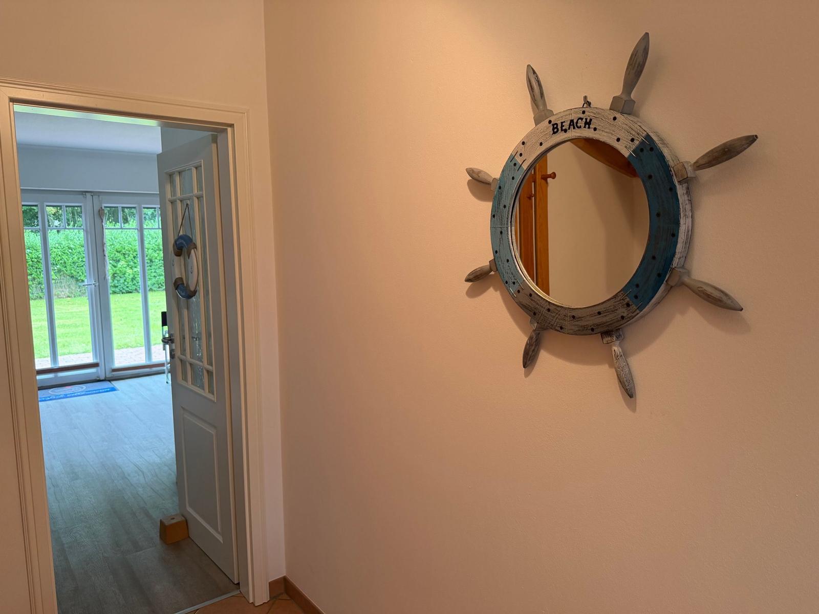 Wandspiegel in Form eines Rudersteuerrads mit Aufschrift BEACH. Tür führt zu einem Raum mit Glasfront.