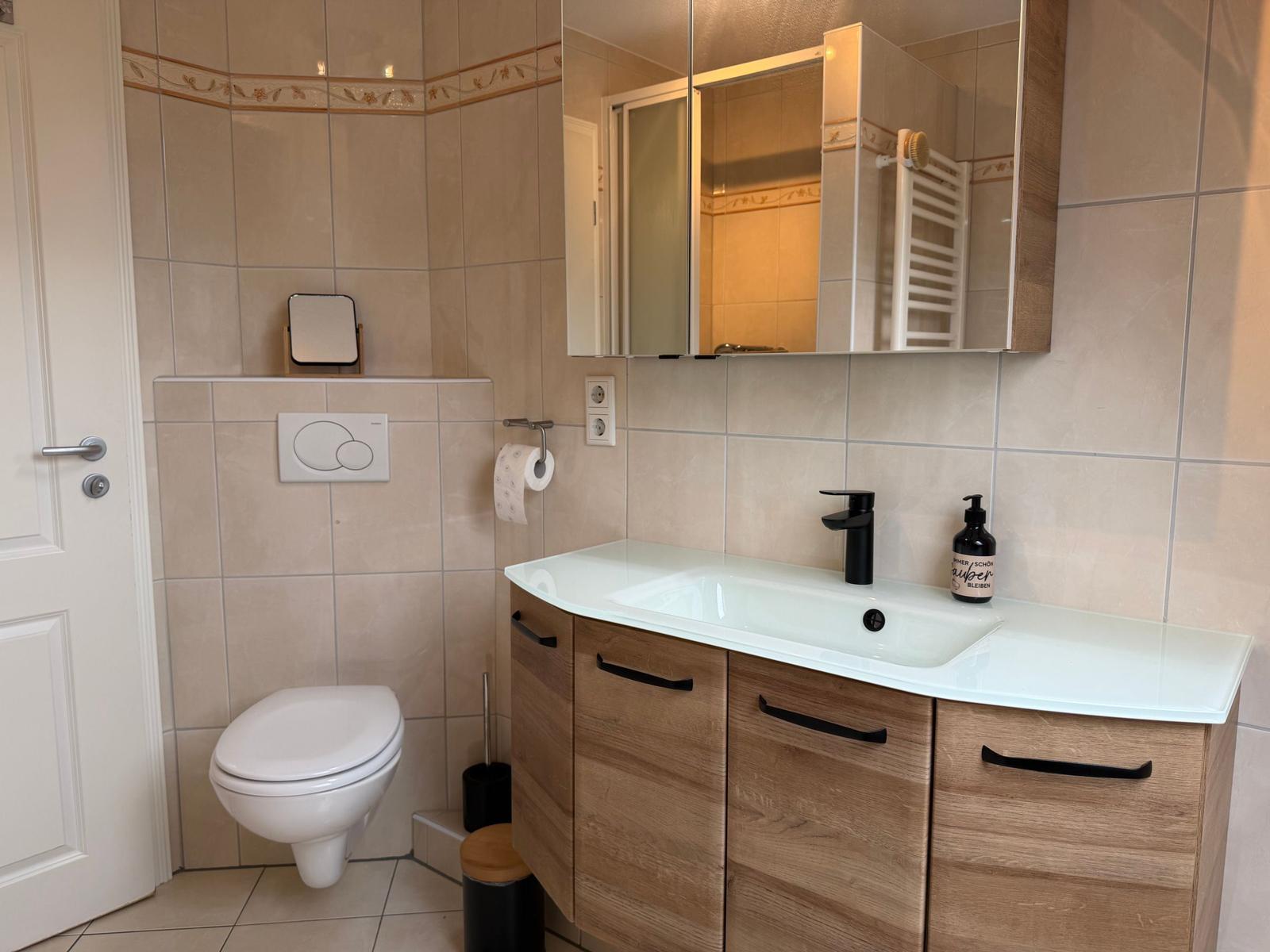 Bad mit Waschtisch, Spiegel und Toilette. Holzschrank und Wandfliesen.