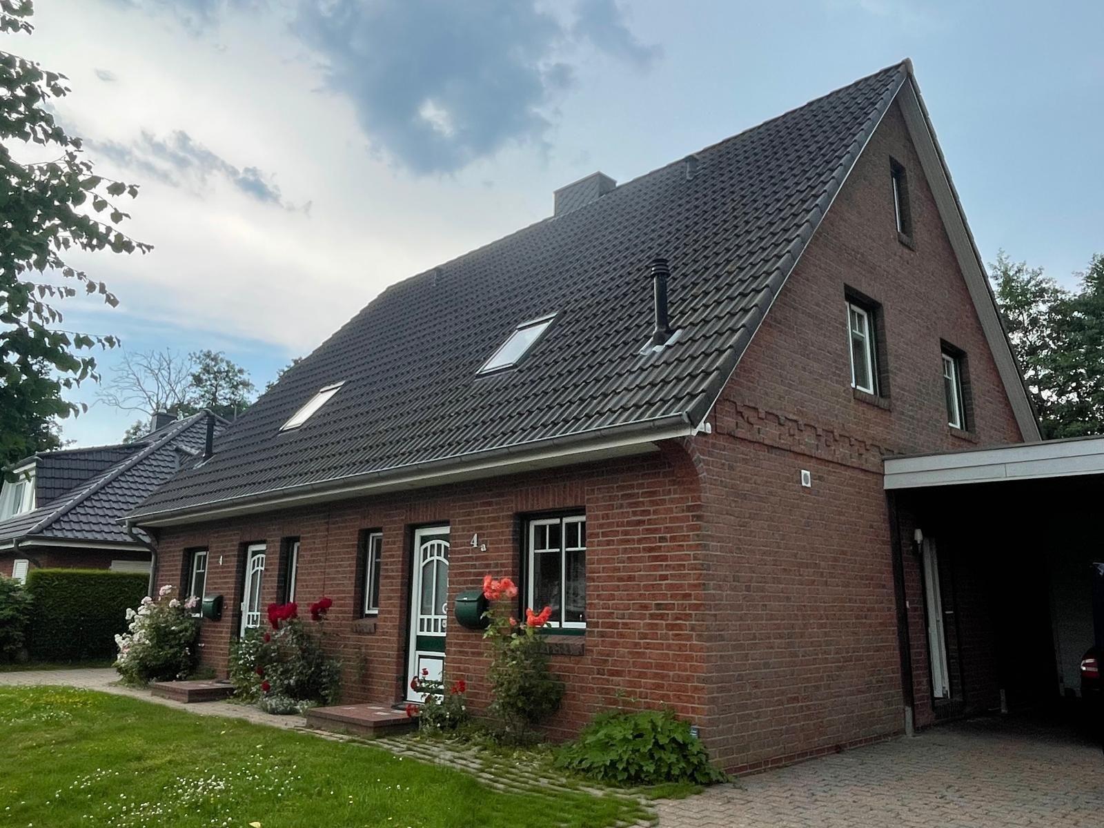 Ein rotes Backsteinhaus mit Dachgauben und Vorgarten. Garage und Blumen sind sichtbar.