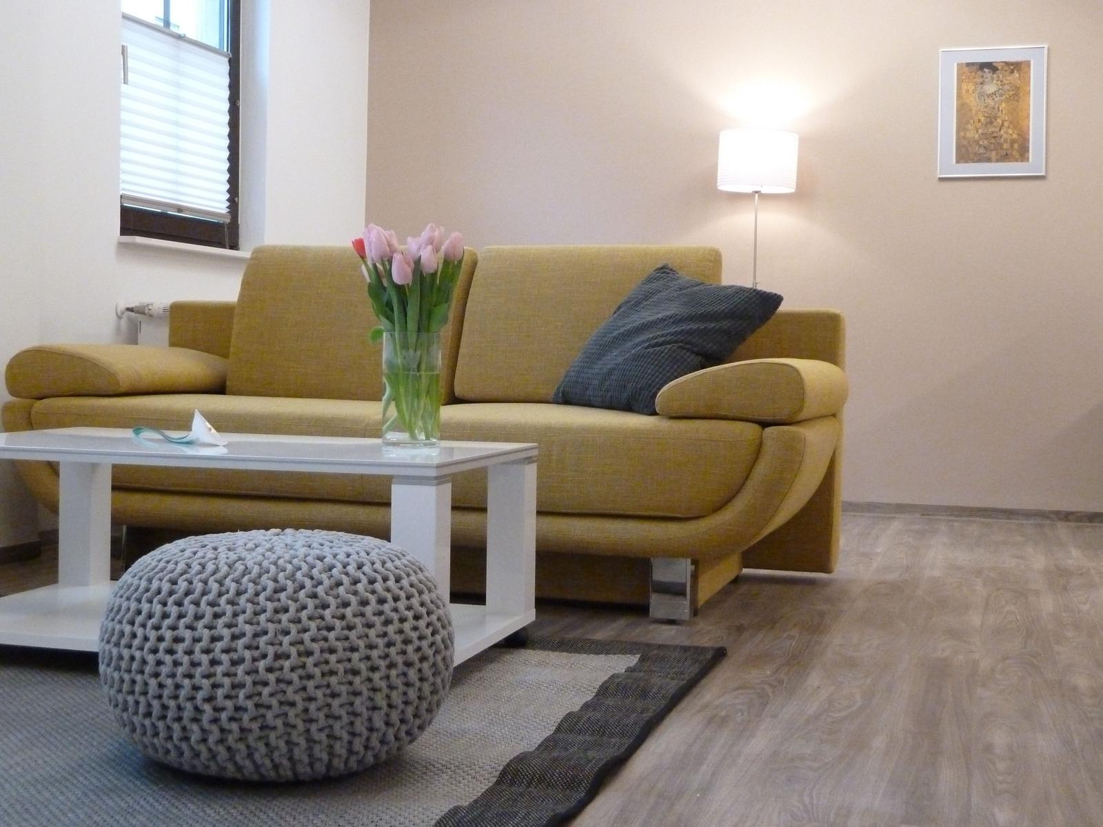 Gelber Sofa mit Kissen, weißer Couchtisch und grauer Hocker. Wandbild und Stehlampe.