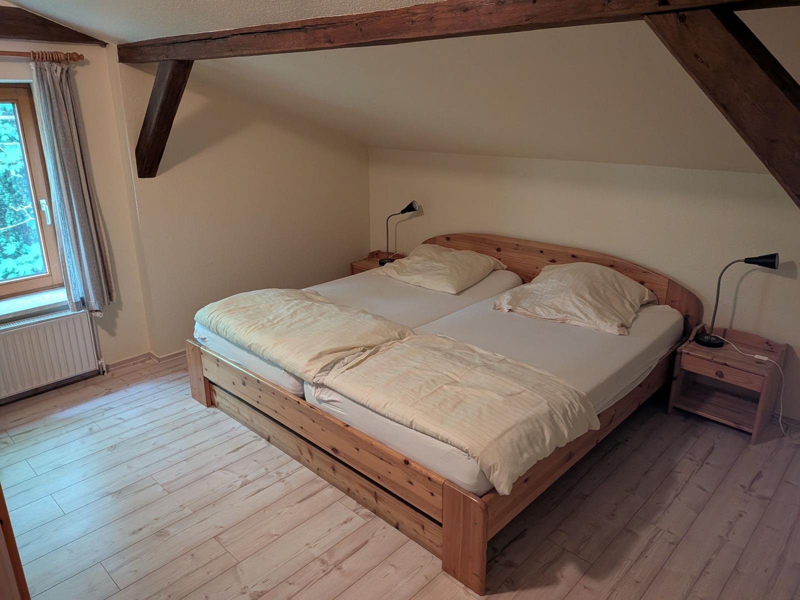 Schlafzimmer mit Doppelbett, Holzboden und Balken. Fenster mit Vorhängen.