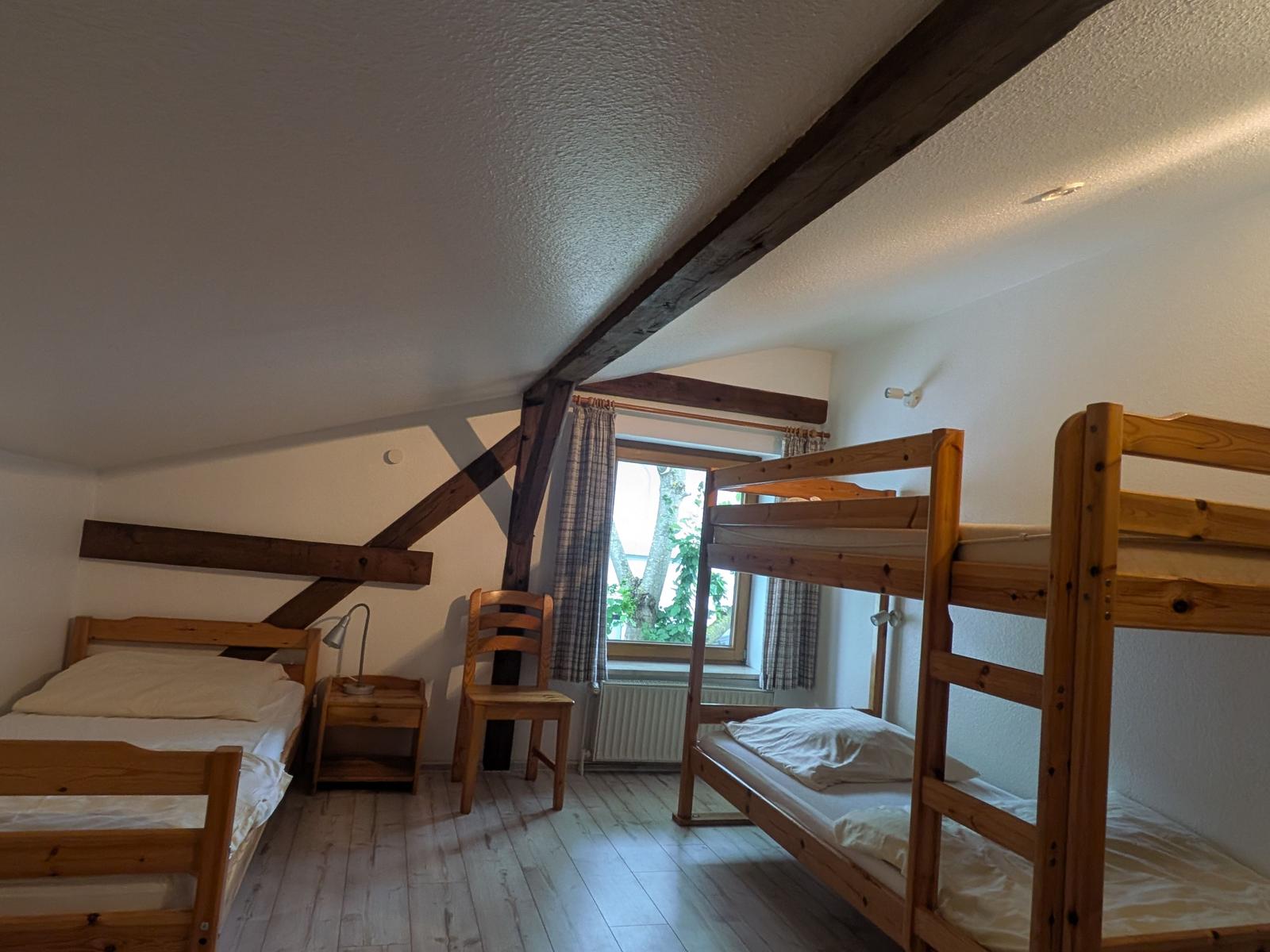 Zimmer mit zwei Etagenbetten und einem Einzelbett, Holzbalken an der Decke, Fenster mit Vorhängen.