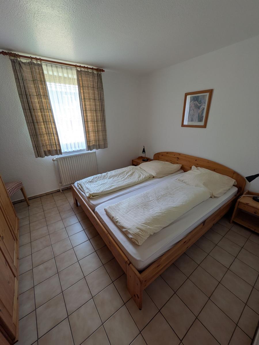 Schlafzimmer mit Doppelbett, Holzbett, Fenster mit Vorhängen und Fliesenboden.