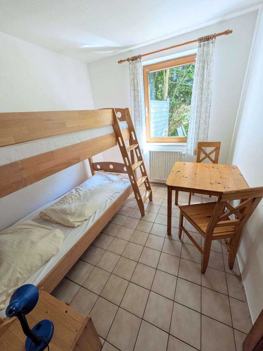 Zimmer mit Holzbett, Tisch und Stühlen. Fenster mit Vorhängen. Fliesenboden.