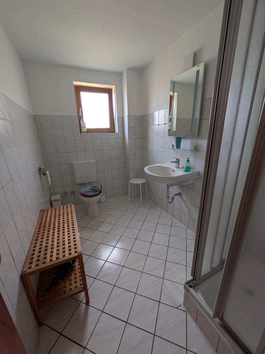 Badezimmer mit Toilette, Waschbecken und Dusche. Holzbank und Fenster sichtbar.