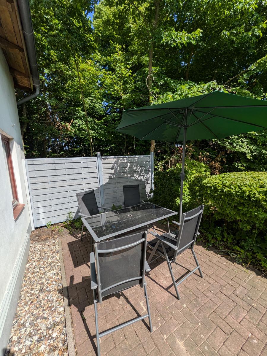 Terrasse mit Tisch, vier Stühlen und grünem Sonnenschirm. Umgeben von Bäumen und Hecke.