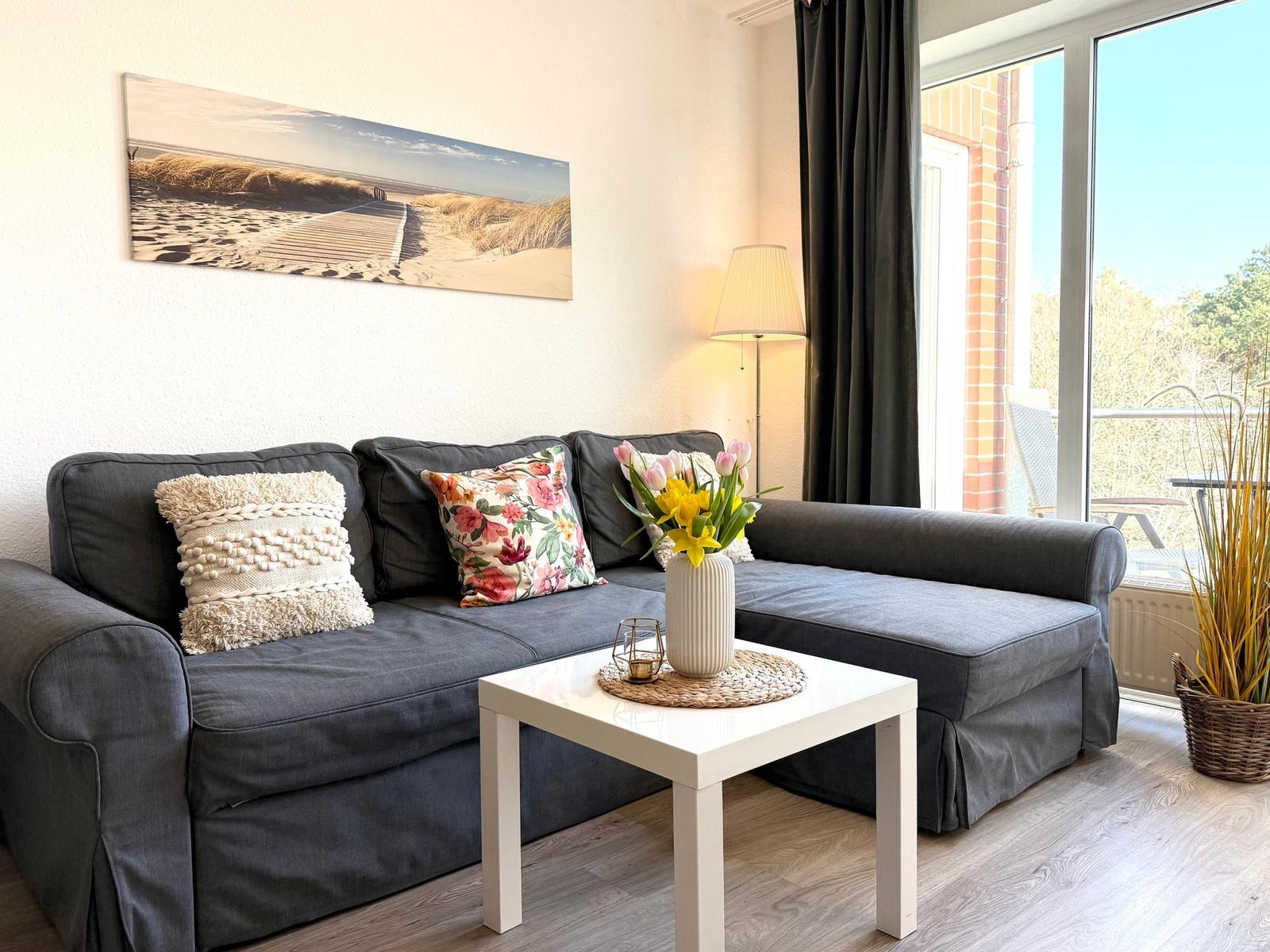 Ein Wohnzimmer mit grauem Sofa, weißen Tisch und Blumen. Große Fenster und Wandbild.