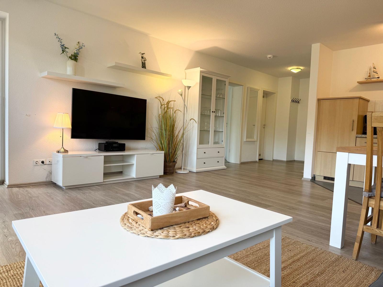 Wohnzimmer mit Fernseher, Couchtisch und Holzboden. Küche und Flur sind sichtbar.