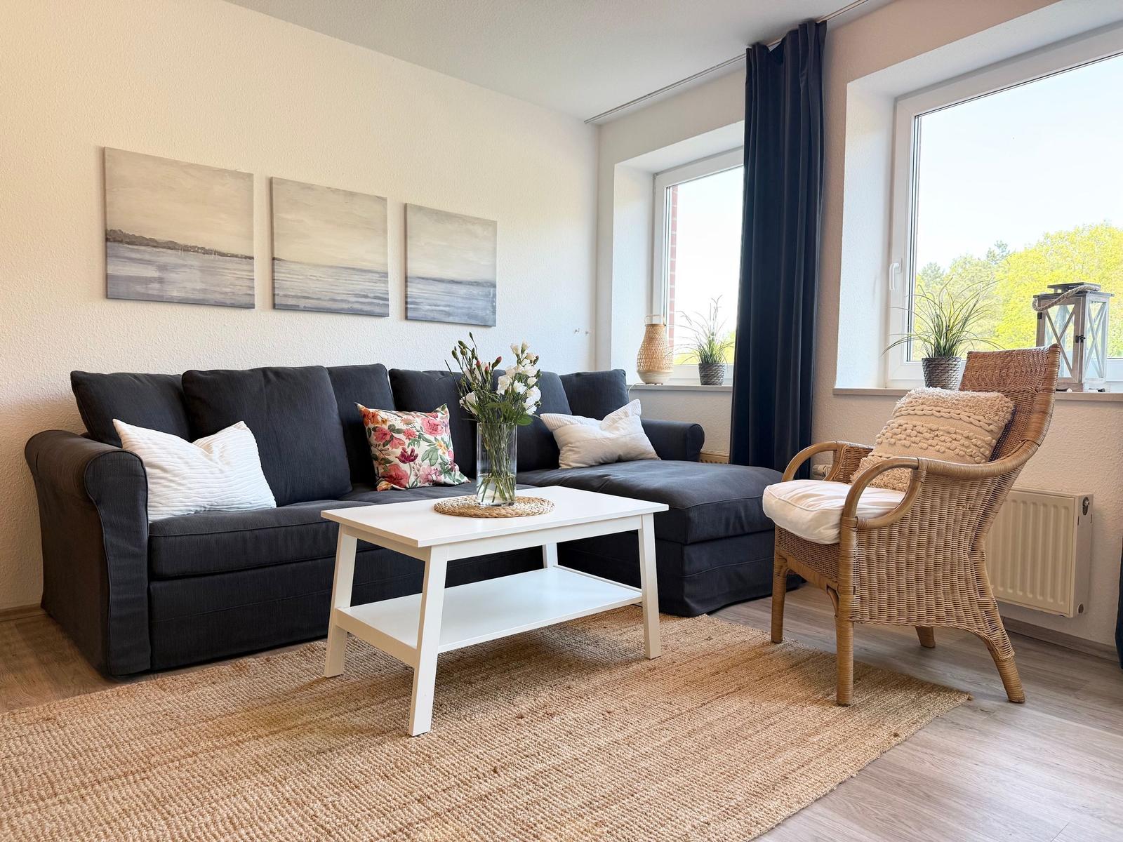 Ein Wohnzimmer mit dunklem Sofa, weißem Tisch und Rattan-Stuhl. An der Wand hängen drei Bilder.