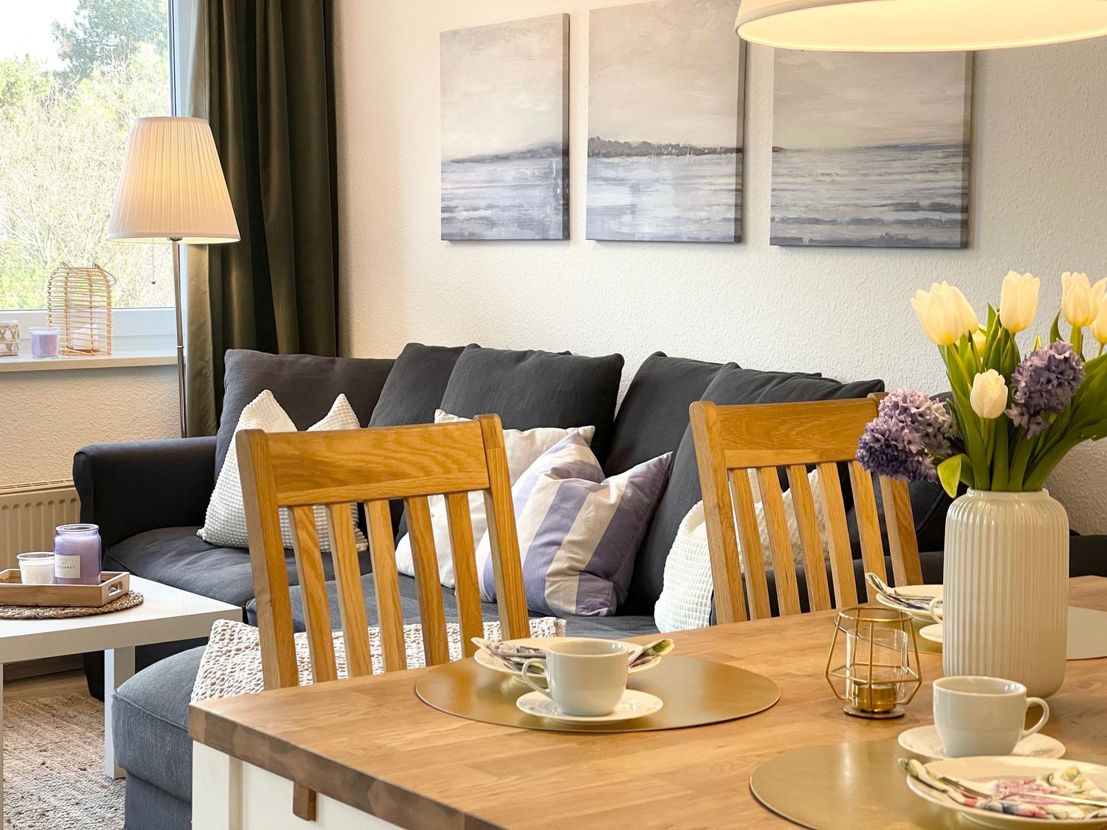 Ein Essbereich mit Holztisch und Stühlen. Im Hintergrund ein Sofa mit Kissen und Wandbildern.