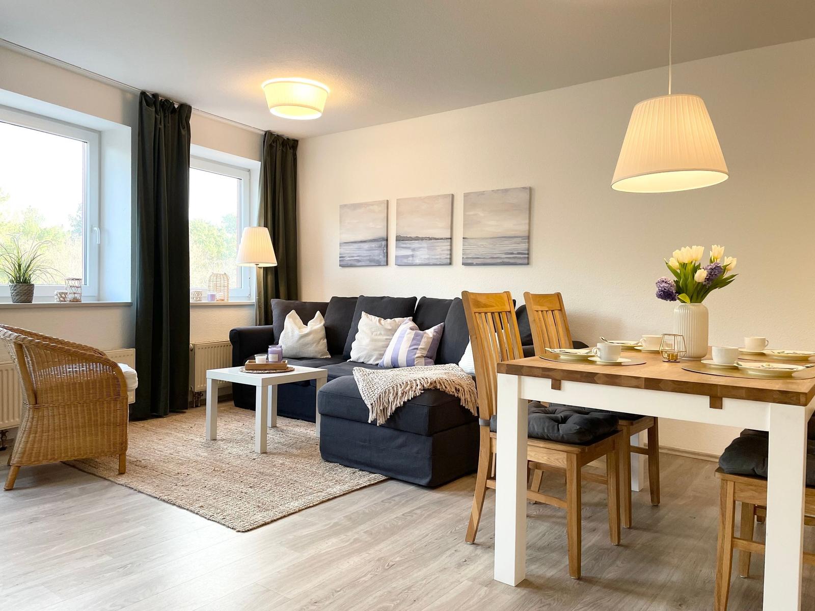 Wohn- und Essbereich mit Sofa, Tisch und Stühlen. Fenster mit Vorhängen und Wandbildern.
