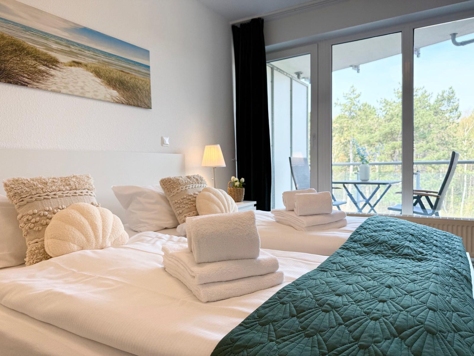 Schlafzimmer mit Balkon, Bett, Kissen und Decke. Blick auf Wald und Tisch mit Stühlen.