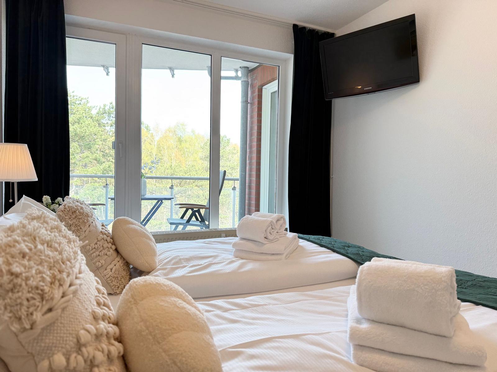 Zimmer mit Doppelbett, TV und Balkon mit Sitzgelegenheit.