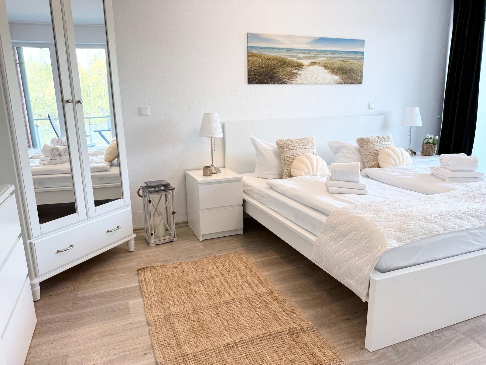 Ein Schlafzimmer mit weißem Bett, Nachttischen und Spiegelschrank. Wandbild zeigt Strand.