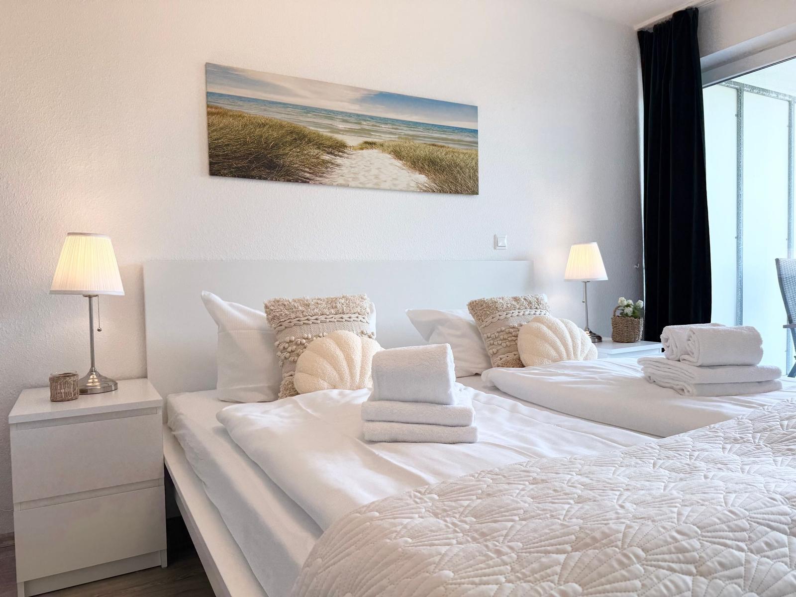 Schlafzimmer mit Doppelbett, zwei Nachttischen und Wandbild. Vorhang und Fenster sichtbar.