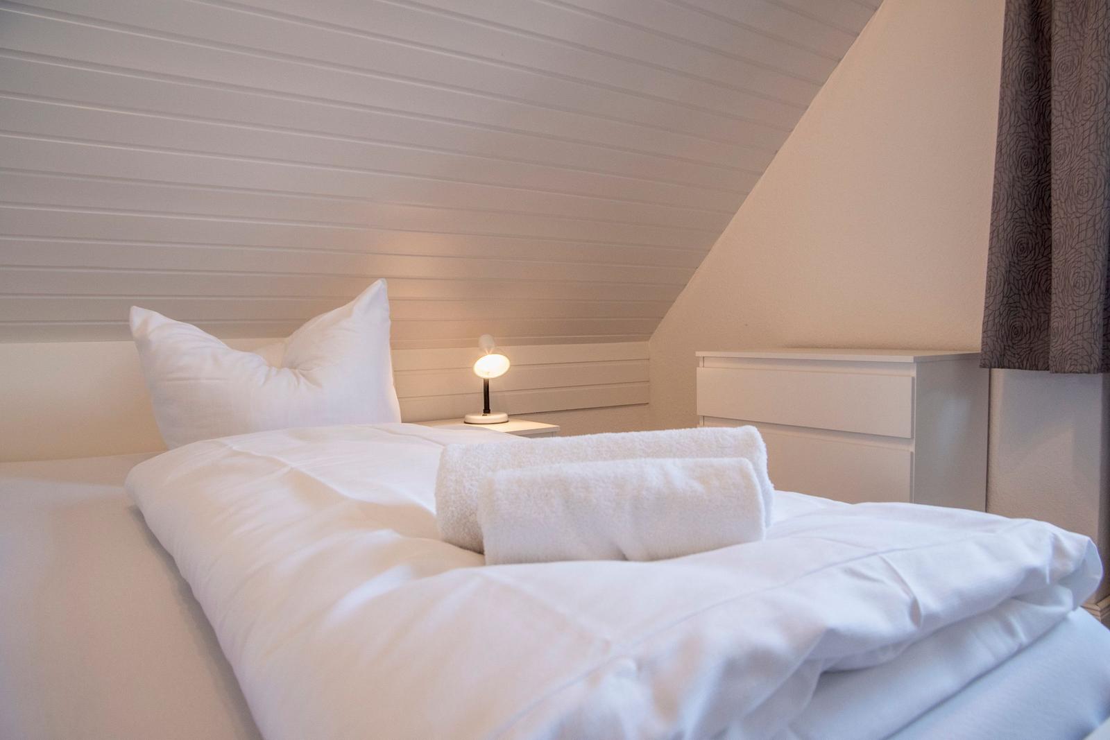Schlafzimmer mit weißem Bett, Nachttisch und Lampen. Dachgeschoss mit schrägen Wänden.