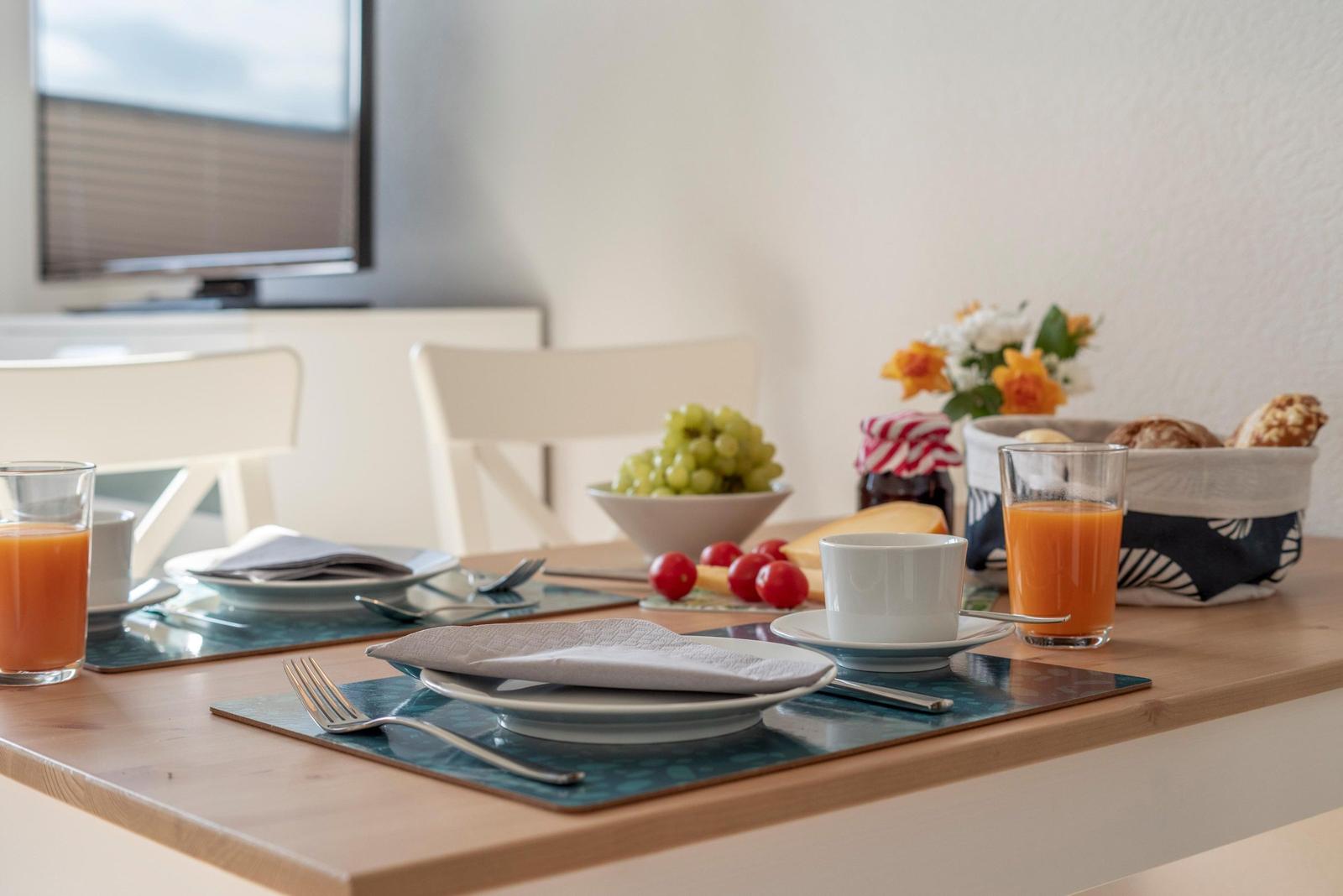 Esstisch mit Obst, Getränken und Tischdecken. Im Hintergrund Fernseher und Blumen.