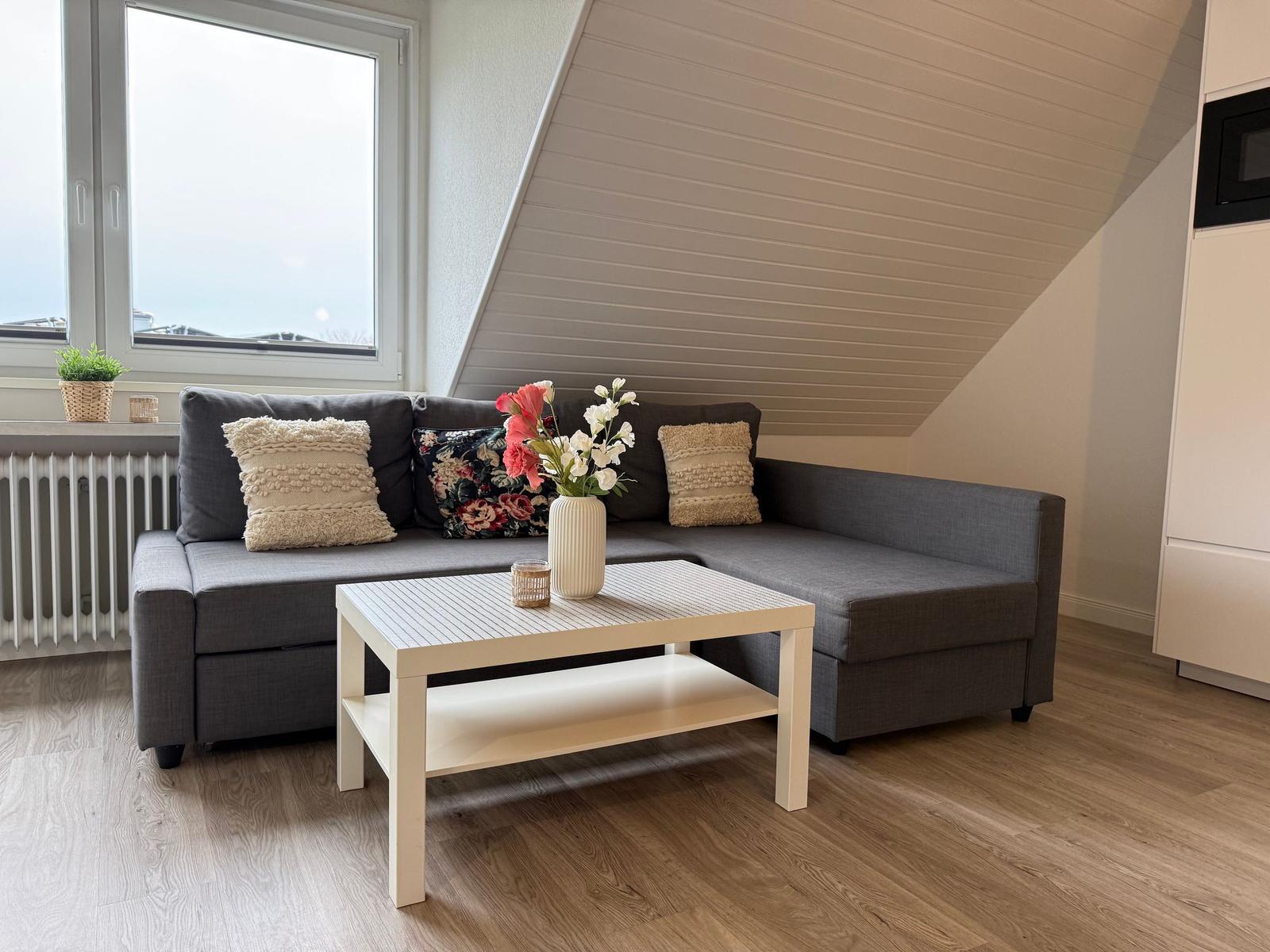 Ein grauer Sofa mit Kissen und Blumen auf einem weißen Tisch. Holzboden und schräge Decke.