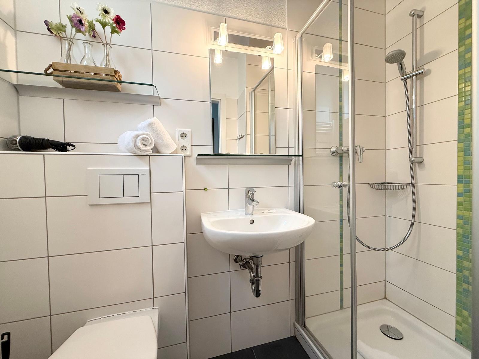 Badezimmer mit Dusche, Waschbecken und Toilette. Weiße Fliesen und grüne Mosaikstreifen.