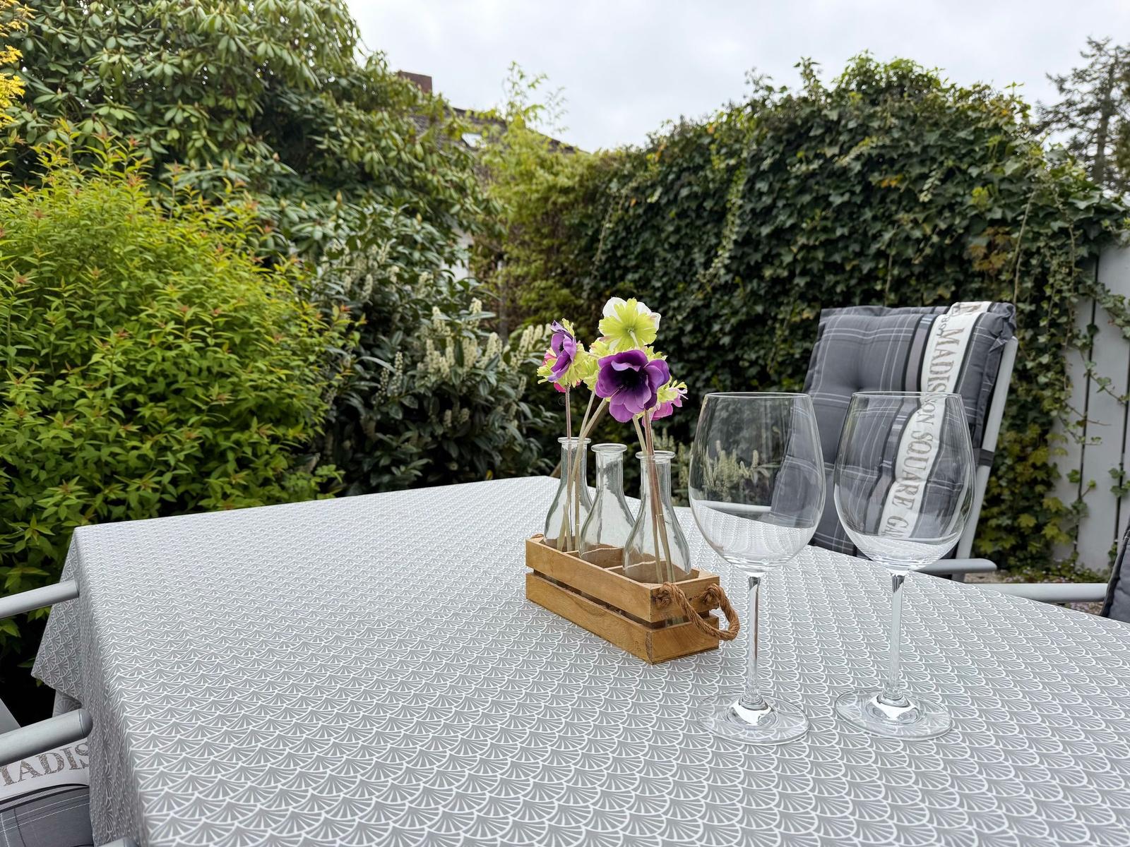 Tisch mit Tischdecke, zwei Weingläsern und Blumen in Flaschen auf einer Terrasse.