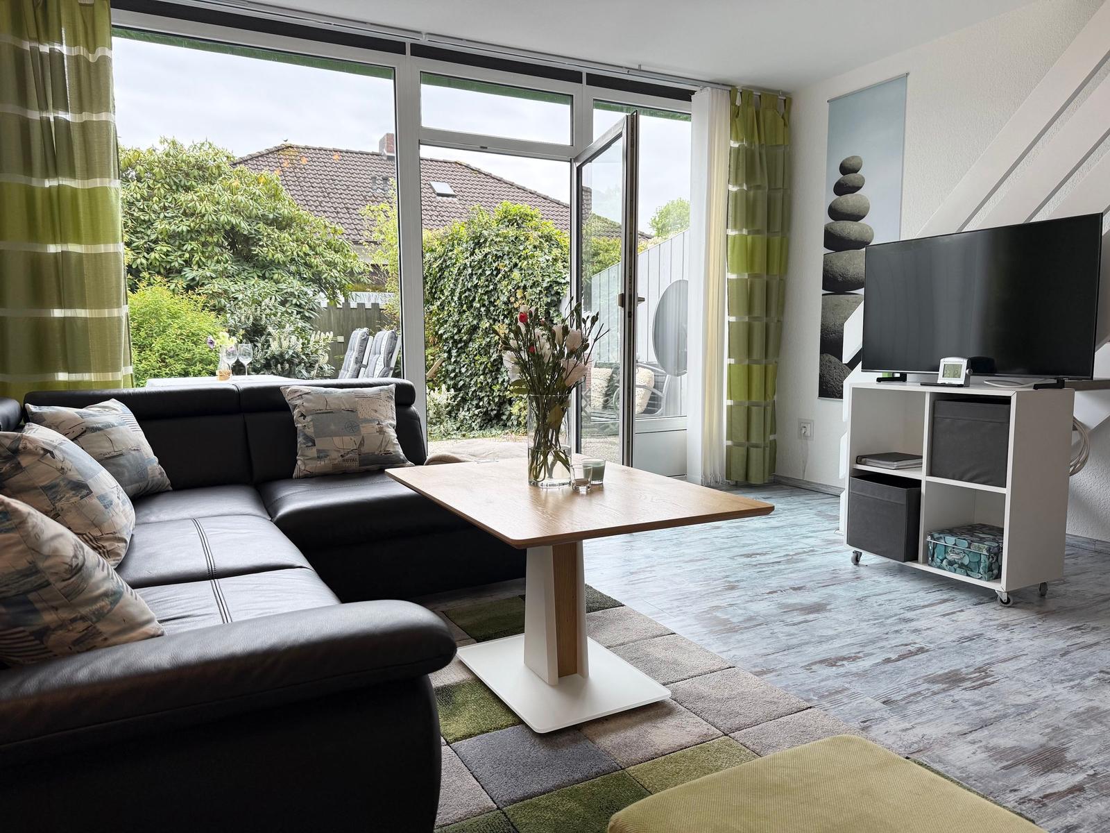 Wohnzimmer mit Sofa, Tisch und TV. Große Fensterfront mit Blick auf Garten.