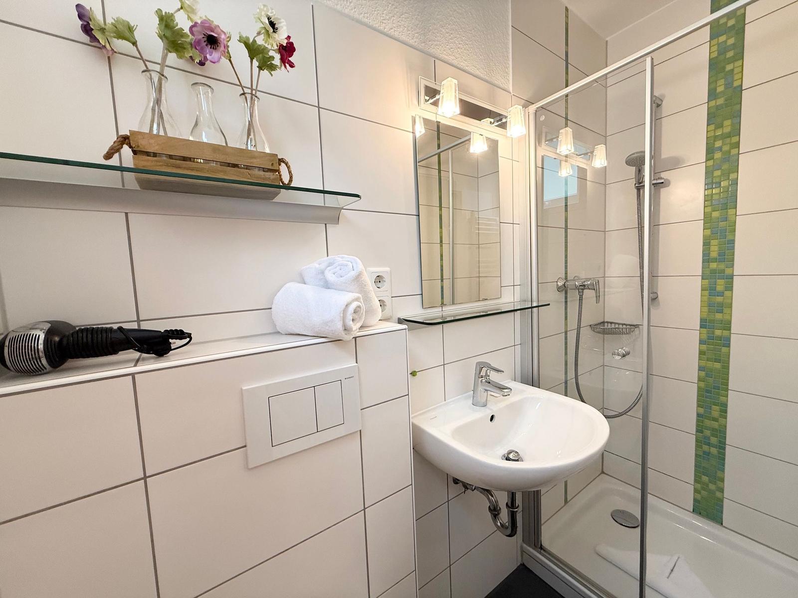 Badezimmer mit Dusche, Waschbecken und Spiegel. Auf Regal Blumen und Handtuch.