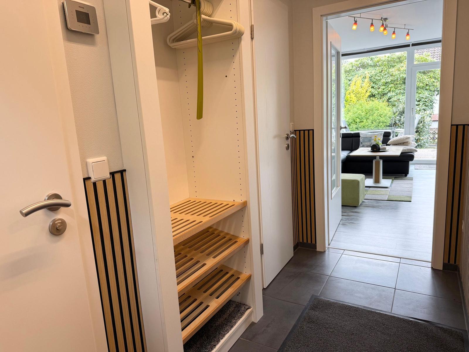 Ein kleiner Schrank mit Holzregalen und Haken. Durch die Tür ist ein Wohnbereich mit Sofa und Tisch sichtbar.
